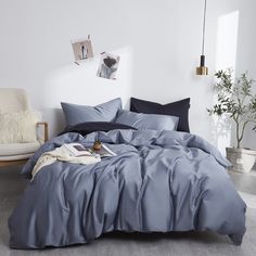 a bed with blue sheets and pillows in a white room next to a chair, potted plant and pictures on the wall