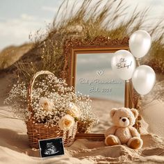 a teddy bear sitting in the sand next to a basket with balloons and a photo frame