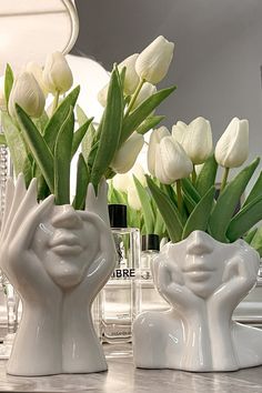 two white vases with flowers in them sitting on a counter next to a mirror