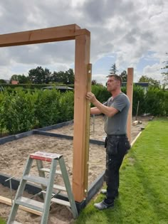 a man standing next to a wooden frame on top of a lush green field with a ladder