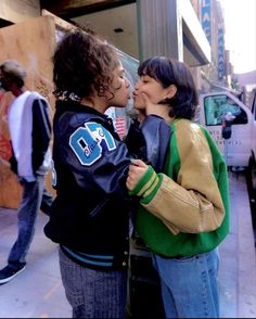 two young women standing next to each other on a sidewalk kissing and smiling at each other