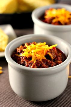 two white bowls filled with chili and cheese on top of a brown place mat next to cornbreads