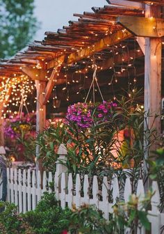 a white picket fence covered in lots of flowers next to a building with lights on it