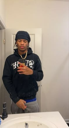 a man standing in front of a bathroom mirror looking at his cell phone while wearing a hat