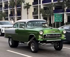 an old green car is driving down the street in front of some cars and palm trees