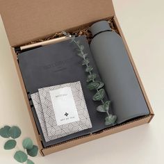 an open box containing a bottle, notebook and some leaves next to it on a white surface