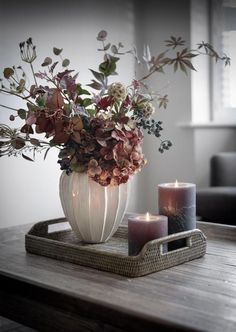 two candles are sitting on a tray with flowers and leaves in the center, along with a vase filled with greenery