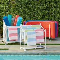 towels are lined up next to the pool in front of some bushes and chairs with blue, pink, orange, and green striped towels hanging on them