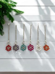 six different colors of beaded flowers hanging from chains on a white wooden surface next to a pine tree