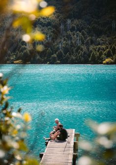 two people sitting on a dock in front of a body of water with trees around them