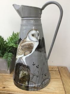 a ceramic pitcher with an owl painted on the front and side, sitting next to a potted plant