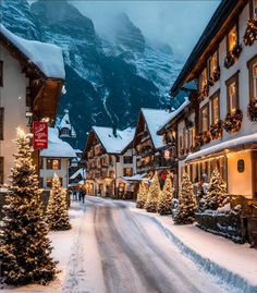 a snowy street with christmas trees and lights on the sidewalks in front of buildings that have snow covered mountains behind them