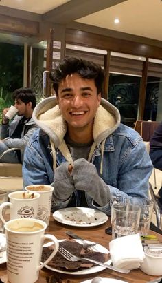 a man sitting at a table with two cups of coffee and plates of food in front of him