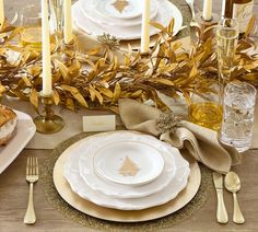 the table is set with white plates and silverware, gold napkins and candles