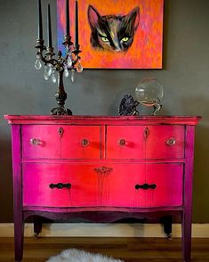 a cat painting on the wall behind a pink dresser with candles and a chandelier