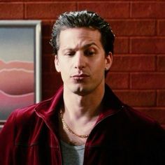 a man standing in front of a brick wall wearing a red shirt and gold necklace