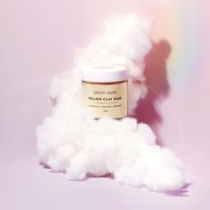 a jar of yellow clay mask sitting on top of white fluffy clouds in front of a rainbow colored background