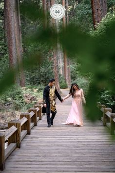 a man and woman holding hands walking across a wooden bridge in the middle of a forest