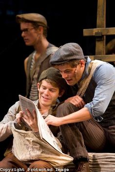 two men sit on the ground and look at a newspaper while another man stands behind them