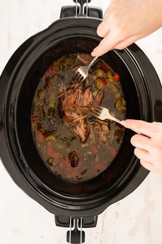 two hands holding a fork and spoon over a slow cooker filled with stew in it