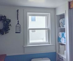 a white toilet sitting under a window next to a shelf filled with blue and white towels