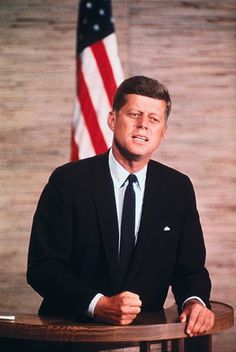 a man in a suit and tie sitting at a table with an american flag behind him