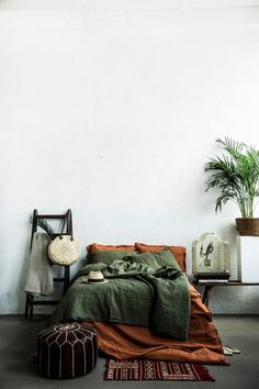 a bed sitting next to a plant in a white room with an orange blanket on top of it