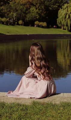 a woman sitting on the grass near water looking at something in the distance with her back turned to the camera