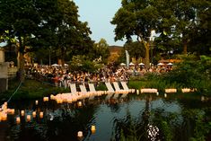 many candles are floating in the water near some trees and lawn chairs with people sitting on them