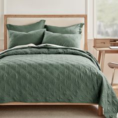 a bed with green bedspread and pillows in a white room next to a desk