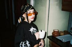 a woman wearing sunglasses and a bandana holding a cup while standing in a kitchen