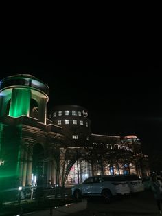 a large building lit up at night with green lights on it's sides and cars parked in front