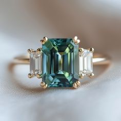 an emerald and diamond ring with three baguets on top, sitting on a white cloth