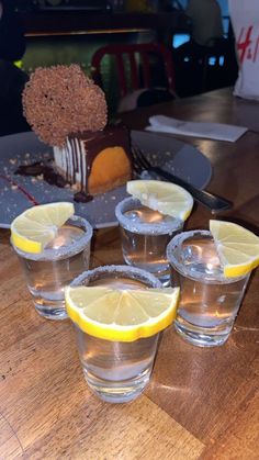 four shot glasses with lemon wedges on a wooden table in front of a cake