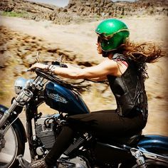 a woman riding on the back of a blue motorcycle down a dirt road with mountains in the background