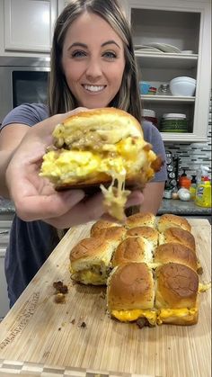 a woman is holding up a sandwich with eggs and cheese on it while standing in front of a cutting board