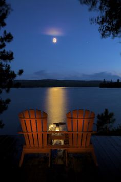 two wooden chairs sitting on top of a pier next to the ocean at night with a full moon in the sky