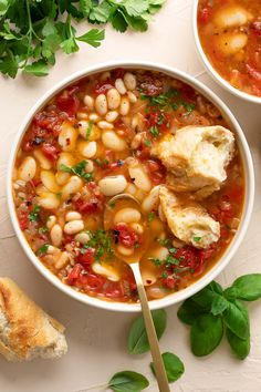 two bowls filled with soup and bread on top of a white table next to fresh herbs