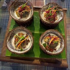 four coconut bowls with meat and vegetables in them on a green tray next to utensils