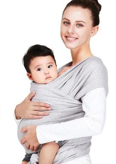 a woman holding a baby wrapped in a grey wrap and smiling at the camera while wearing a white shirt