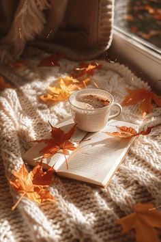 a cup of coffee on top of an open book with autumn leaves around it and a window sill in the background