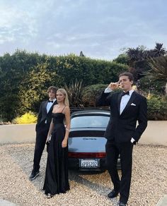 two people in tuxedos are standing next to a car and one person is taking a photo