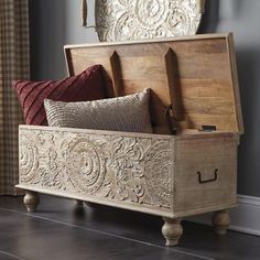 a wooden trunk with decorative carvings on the front and sides, sitting in a living room next to a window