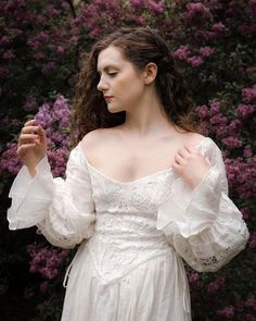 a woman wearing a white dress standing in front of purple flowers