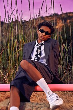 a young man wearing sunglasses and a suit sitting on a bench in front of tall grass