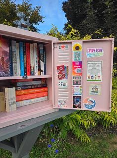 a pink book case filled with lots of books on top of a wooden bench next to trees