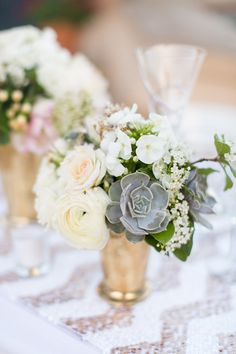 two vases filled with white flowers and greenery on top of a lace table cloth