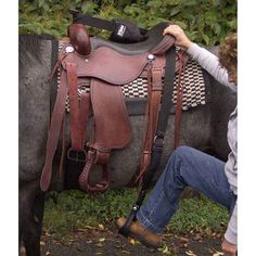 a woman kneeling down next to a brown horse with a saddle on it's back