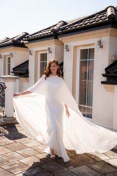 a woman wearing a white dress and cape standing in front of a house with her hands on her hips