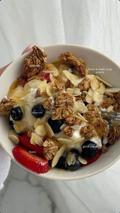 a bowl filled with granola and fruit on top of a white tablecloth next to a person holding a fork